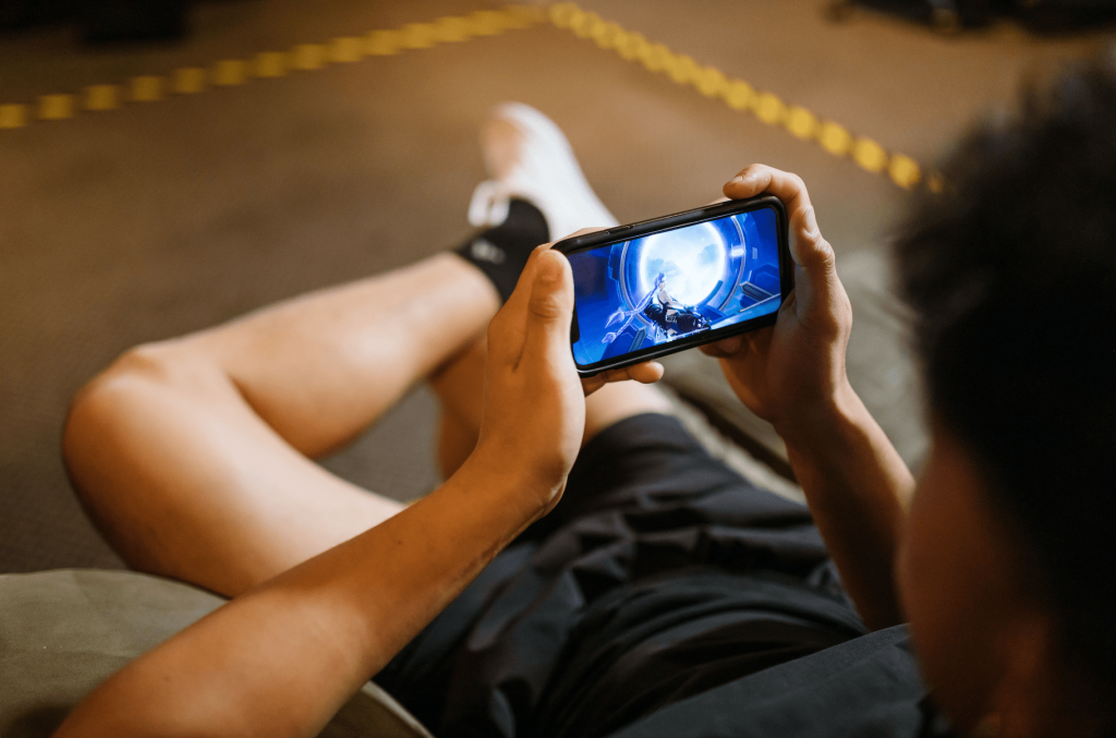 A person is seen reclining on a couch while playing a game on their smartphone. The individual is holding the phone with both hands, and the screen displays a futuristic, action-packed game scene featuring a character in a high-tech environment. The person's legs are stretched out, wearing casual athletic shorts and sneakers. The background is slightly blurred, focusing the attention on the gaming activity. The setting appears relaxed and comfortable, emphasizing a leisure moment spent on mobile gaming.