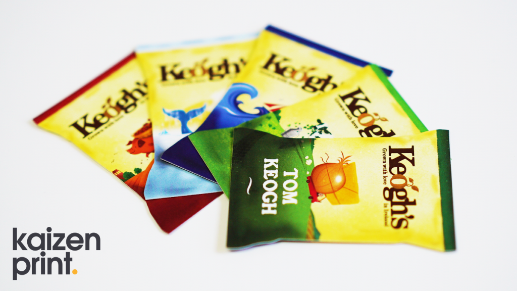 A close-up image of several colorful snack bags from Keogh's, fanned out on a white background (traditional ad). The packaging displays different vibrant designs, each representing a unique flavor. The bag in the front has a green theme and reads "TOM KEOGH." The other bags behind it feature a mix of yellow, blue, and red colors with various illustrations. The Keogh's logo, "Grown with love in Ireland," is prominently displayed on each bag. The Kaizen Print logo is in the bottom left corner of the image, indicating the packaging was printed by Kaizen Print.