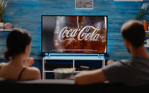 A couple is sitting on a couch watching a Coca-Cola commercial on a flat-screen TV (traditional ad). The screen displays a close-up of a Coca-Cola glass with the Coca-Cola logo prominently featured. The room has a cozy atmosphere with a blue wall in the background and some decorative items on shelves. The couple is seen from behind, focusing on the TV, with a bowl of snacks placed on a table in front of them.