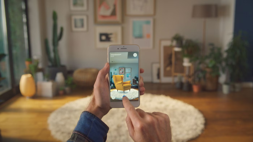A person using a smartphone to visualize how a piece of furniture, specifically a yellow armchair, would look in their living room through an augmented reality (AR) app. The image demonstrates the integration of AR technology in home decor and furniture shopping, allowing users to virtually place and view furniture in their home environment before making a purchase (AR).