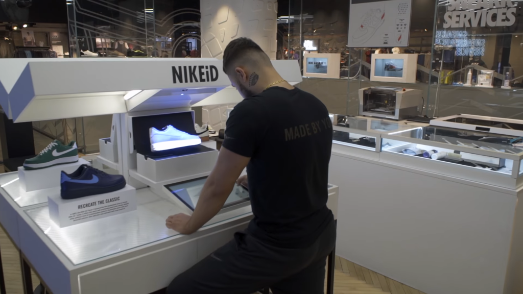 A customer interacts with Nike's VR store experience at a NIKEiD customization station. The setup features digital screens and interactive touch displays, allowing the customer to design and visualize their own custom sneakers in real-time. This immersive technology provides a unique and engaging way for shoppers to personalize their purchases, enhancing the overall shopping experience.
