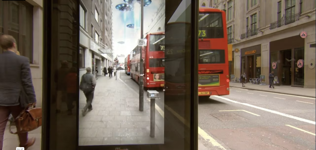 A bus shelter in London with an augmented reality (AR) display. The AR screen shows a surprising and imaginative scene where flying saucers are hovering over the city street, blending seamlessly with the real world surroundings. This interactive ad campaign is designed to capture the attention of passersby by integrating unexpected and engaging digital content with the physical environment.