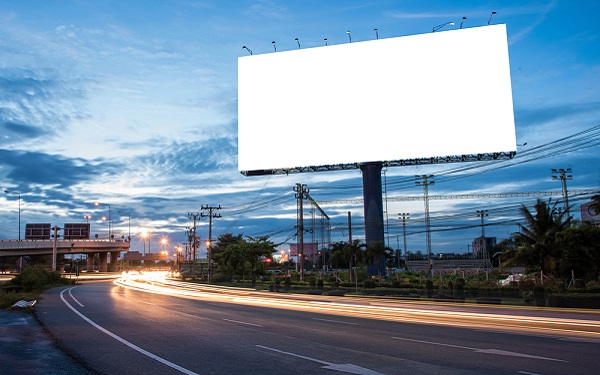 A large blank billboard stands beside a highway, illuminated against the twilight sky. The empty billboard is ready for advertising content, with a few lights on top aimed at the display area. Below, the highway curves with light trails from passing vehicles, indicating movement and activity. The backdrop features a mix of urban infrastructure and vegetation, with streetlights and power lines visible in the distance.