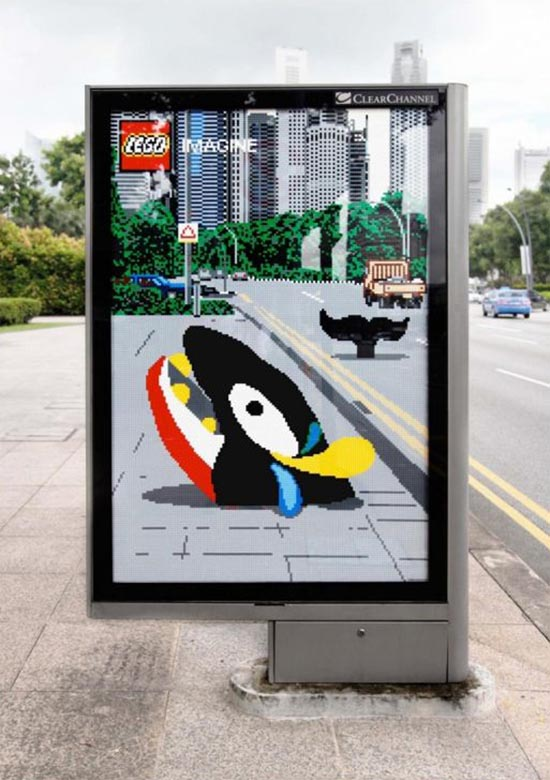 A creative bus stop advertisement features a LEGO-themed display. The ad depicts a pixelated scene of a road with buildings in the background, created entirely with LEGO bricks. In the center of the image, a colorful, cartoonish character appears to be breaking through the pavement, adding a playful and imaginative touch. The top left corner of the ad shows the LEGO logo with the word "IMAGINE" next to it. The ad is designed to inspire creativity and highlight the versatility of LEGO bricks. The surrounding environment includes a city street with vehicles and greenery.