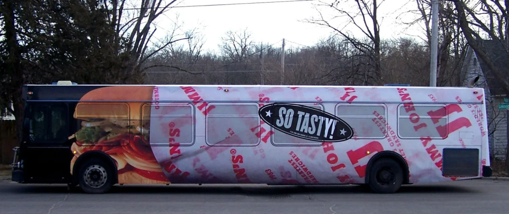 A city bus wrapped in a full-body advertisement for a fast-food brand. The ad features a large, mouth-watering sandwich image towards the front of the bus, with the rest of the bus wrapped in branded sandwich paper displaying the text "Jimmy John's" repeatedly. In the center of the bus, the words "SO TASTY!" are prominently displayed inside a black oval. The bus is parked on a street with bare trees and houses in the background, suggesting a winter setting.