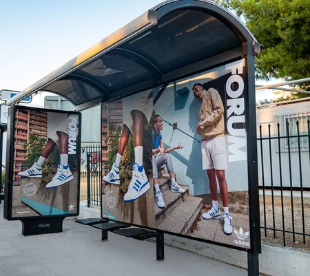 A bus shelter advertisement (OOH) featuring a vibrant Adidas campaign for their "Forum" sneakers. The ad showcases two large posters with stylish images of people wearing the Adidas Forum sneakers. One poster shows a close-up of legs wearing the sneakers, while the other features two individuals casually interacting and showing off their shoes. The word "FORUM" is prominently displayed vertically on the right side of the shelter. The setting includes an urban backdrop with trees, a fence, and buildings in the background, creating a modern and energetic atmosphere.