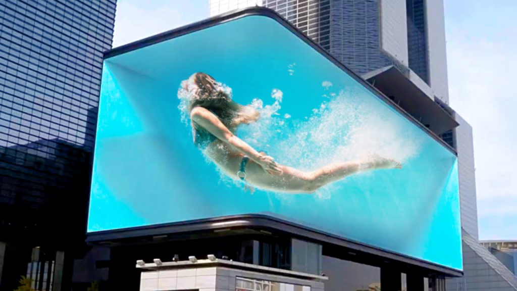 A large digital billboard (DOOH) on the side of a building, displaying an advertisement that creates the illusion of a person swimming underwater. The dynamic, 3D-like visual shows a woman in mid-swim, with bubbles and water splashes enhancing the realistic effect. The surrounding area includes modern high-rise buildings, contributing to the urban and innovative atmosphere of the digital advertisement.