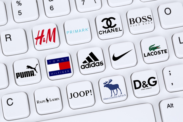 Close-up of a white keyboard featuring keys adorned with logos of popular fashion brands such as H&M, Primark, Chanel, Boss, Puma, Tommy Hilfiger, Adidas, Nike, Lacoste, Ralph Lauren, JOOP!, Abercrombie & Fitch, and D&G. This image symbolizes the integration of fashion brands in the digital and online space