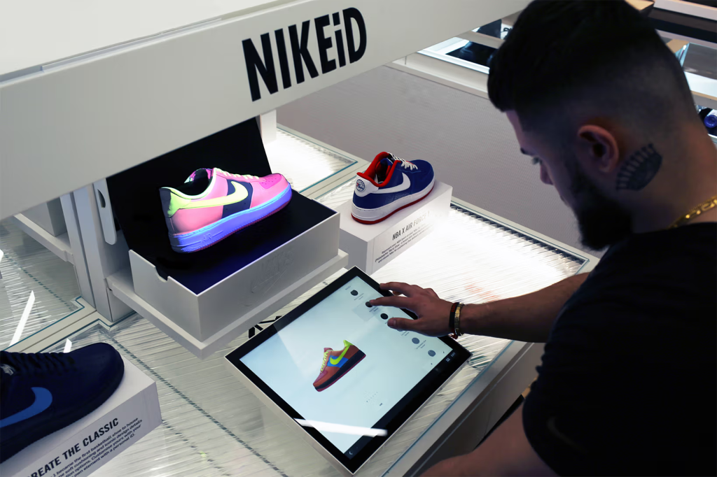 A person using a tablet to customize a pair of Nike sneakers in a NIKEiD store. The display shows various shoe models and customization options, highlighting the interactive and personalized shopping experience.