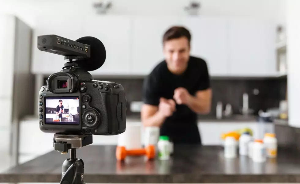 A person is seen recording a video in a kitchen. The camera is set up on a tripod, and the person is focused on explaining or showcasing various products, which are placed on a countertop in front of them. The background is slightly blurred, emphasizing the person and the camera in the foreground.