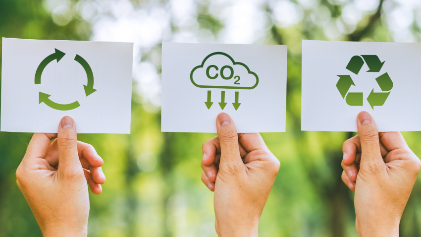 Three hands hold up signs against a backdrop of greenery. The signs feature green icons related to sustainability: the first shows a circular arrow symbolizing the cycle of reuse, the second depicts a cloud with "CO2" and downward arrows representing carbon reduction, and the third displays the recycling symbol. This imagery underscores themes of environmental conservation and eco-friendly practices.