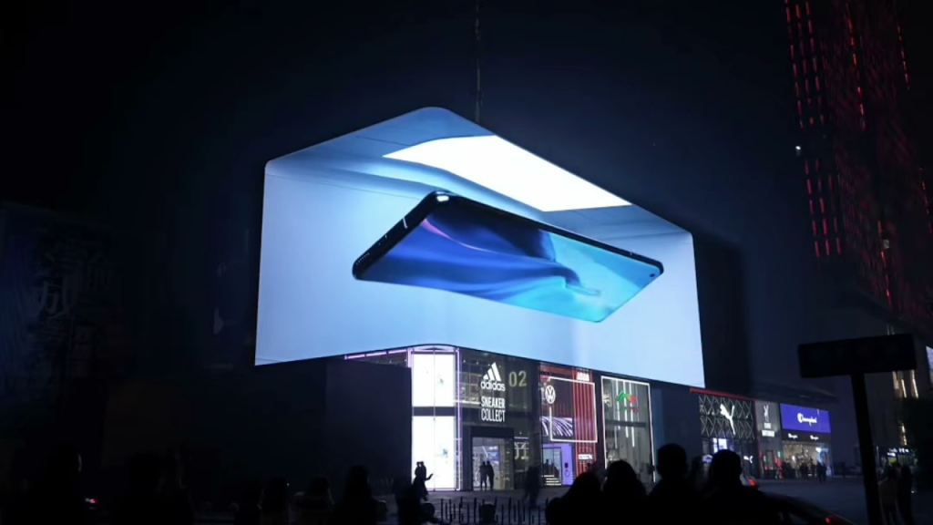 A 3D billboard (DOOH) in a bustling city at night displays a vivid and realistic image of a smartphone. The phone appears to float and rotate in mid-air, captivating the attention of pedestrians below. This innovative use of 3D advertising technology creates a dynamic and engaging visual experience for viewers.