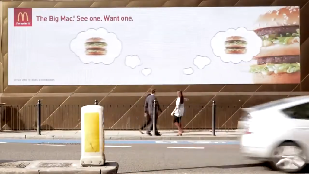 A McDonald's digital billboard (DOOH) displays an advertisement for the Big Mac with the slogan "The Big Mac. See one. Want one." The ad features two images of the Big Mac in thought bubbles, suggesting that seeing the burger will make viewers crave it. In the foreground, a car speeds past, and two pedestrians walk by, highlighting the ad's location on a busy city street.