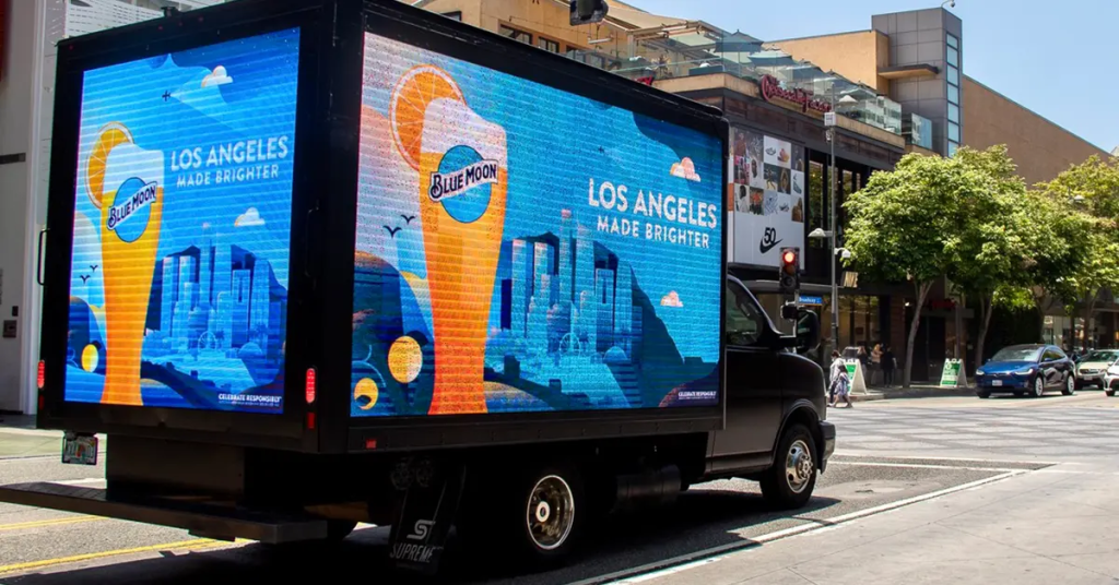 A mobile digital billboard truck (DOOH) displaying an advertisement for Blue Moon beer. The ad features a bright and colorful graphic with a large glass of Blue Moon beer and the text "LOS ANGELES MADE BRIGHTER." The digital display showcases a cityscape of Los Angeles with a clear blue sky, creating a vibrant and inviting scene. The truck is driving through a busy urban area with shops and buildings in the background, emphasizing the dynamic nature of the advertisement.