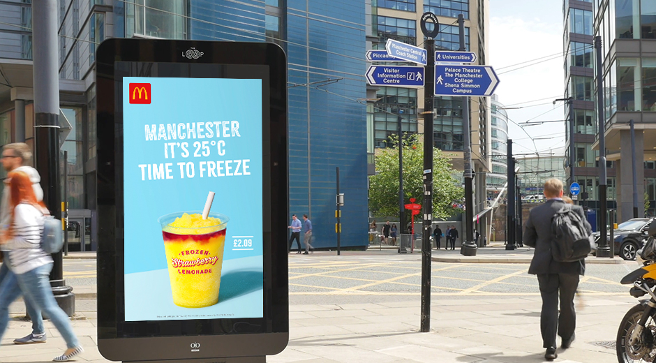 An outdoor digital billboard (DOOH) in Manchester displays an advertisement for McDonald's. The ad highlights the current temperature, stating "MANCHESTER IT'S 25°C TIME TO FREEZE," and promotes a Frozen Strawberry Lemonade priced at £2.09. The vibrant image of the drink on a light blue background catches attention. In the surrounding area, people are walking, and the backdrop includes modern buildings and street signs pointing to various city locations, adding to the urban atmosphere.