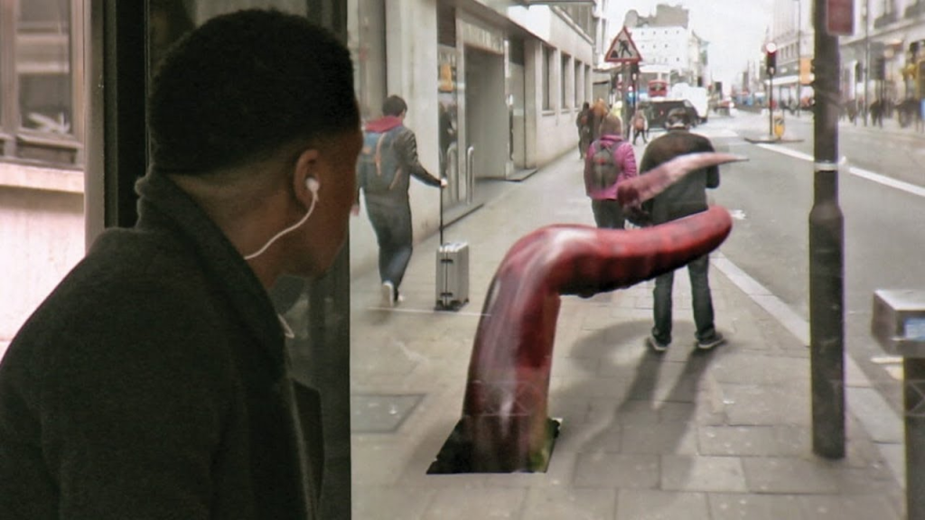 A man with earphones looks out from a bus stop, seemingly startled, as a large, realistic tentacle emerges from a manhole on the street. The scene creates a surreal and dramatic visual effect, making it appear as if a giant creature is invading the city. Pedestrians in the background continue with their day, unaware of the visual spectacle, enhancing the contrast between the normal urban environment and the fantastical element of the tentacle. This image is an example of augmented reality used in advertising to capture attention and create a memorable experience.






