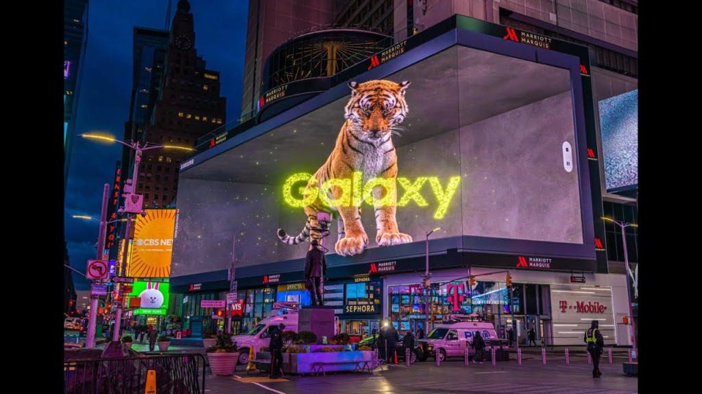 A large 3D billboard (DOOH) in a bustling city, possibly Times Square, displays a striking image of a tiger with the word "Galaxy" illuminated in bright yellow beside it. The billboard is designed to create an immersive 3D effect, making it appear as if the tiger is walking out of the screen. The surrounding area is filled with other advertisements, shops, and people, typical of a vibrant cityscape. This impressive display showcases the advanced use of digital advertising to captivate and engage viewers with realistic and dynamic visual effects.