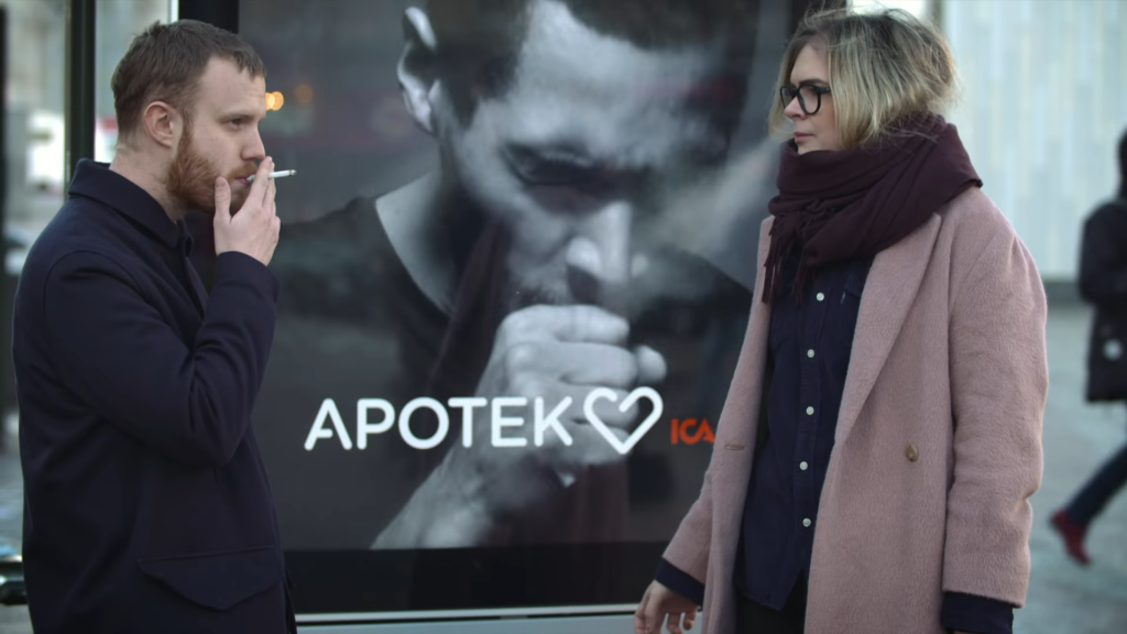 A man is smoking a cigarette next to a woman on a city street. Behind them, a digital billboard from Apotek, a pharmacy, displays an image of a man coughing, indicating the adverse effects of smoking. The billboard's text reads "APOTEK" with a heart symbol, followed by "ICA," suggesting a public health message or anti-smoking campaign.